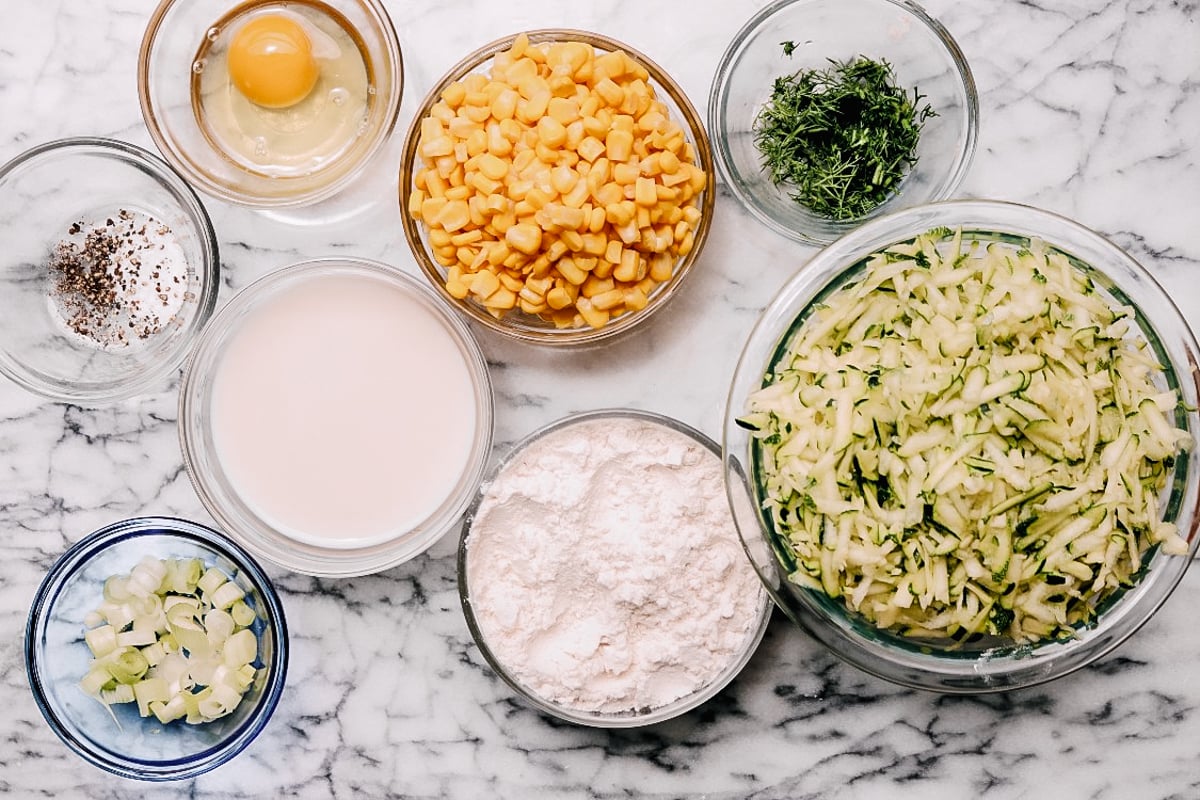 Various ingredients for Zucchini and Corn Fritters are arranged in glass bowls on a marble surface, including an egg, corn, chopped herbs, grated zucchini, flour, sliced green onions, milk, and a mix of salt and pepper.