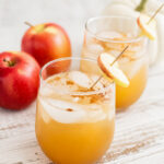 Two glasses of iced apple cider with cinnamon, garnished with an apple slice, sit on a white wooden surface next to two red apples and a white pumpkin, creating the perfect scene for enjoying a refreshing Apple Gin Cocktail.
