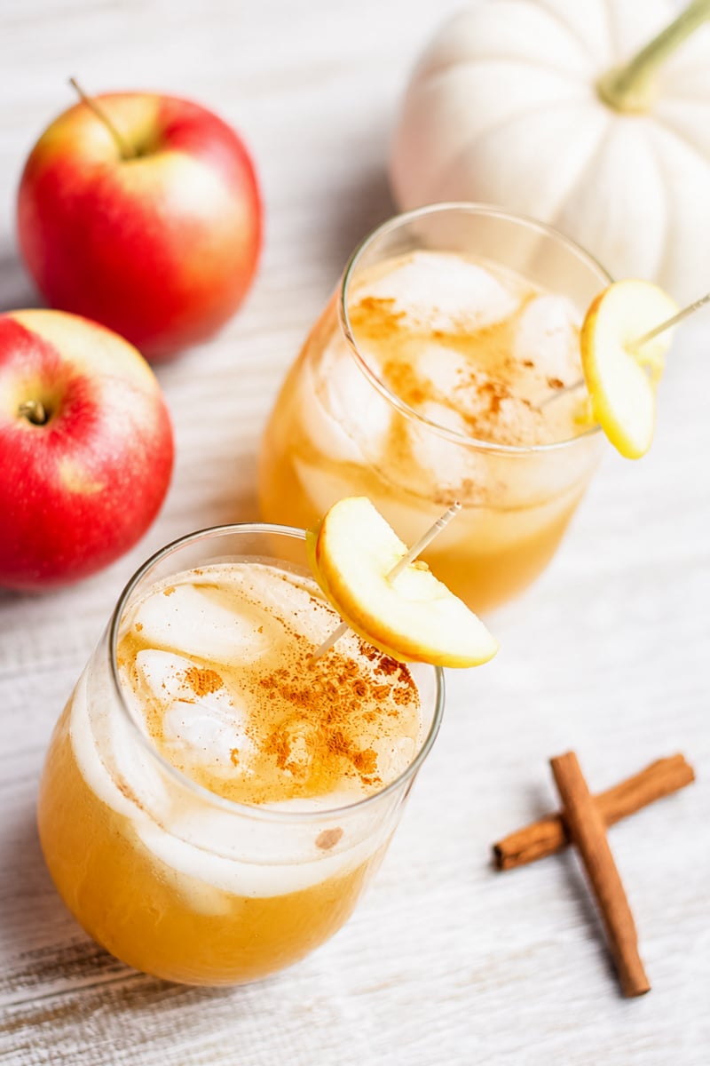 Two glasses of iced apple cider, reminiscent of a refreshing Apple Gin Cocktail, garnished with apple slices and sprinkled cinnamon, with fresh apples, a white pumpkin, and cinnamon sticks on a light wooden surface.