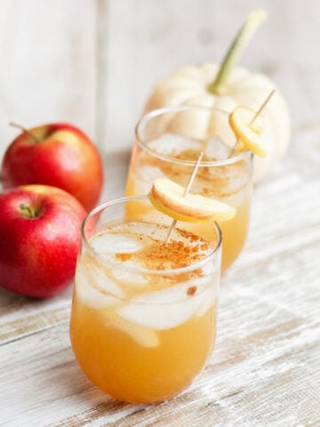 Two glasses of iced apple cider, reminiscent of an Apple Gin Cocktail, are garnished with fresh apple slices and cinnamon on a white wooden surface, with two red apples and a small white pumpkin in the background.