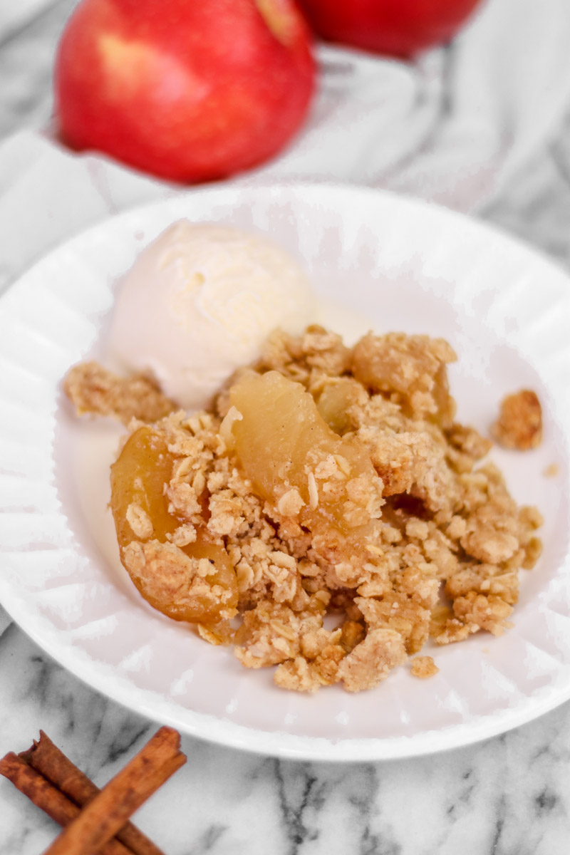 overhead image of cinnamon apple crisp on a white plate and apples in background.