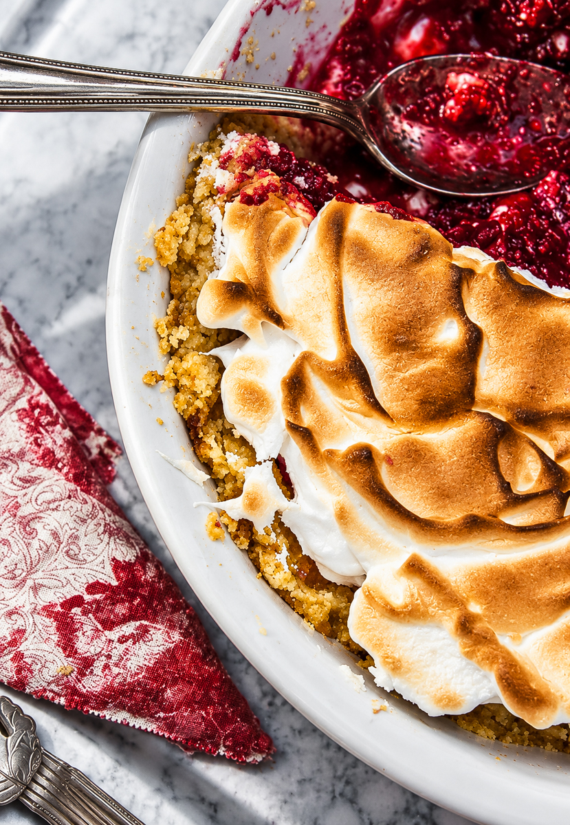 A serving dish containing a cranberry meringue pie with toasted topping and a silver serving spoon resting inside.