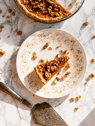 A slice of Amaretti Almond pumpkin pie with nut crumble topping on a floral patterned plate, with the rest of the pie in a glass dish nearby and a pie server on a marble surface.