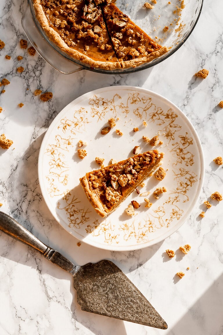 A slice of Amaretti Almond pumpkin pie with nut crumble topping on a floral patterned plate, with the rest of the pie in a glass dish nearby and a pie server on a marble surface.