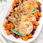 Three cooked pork chops garnished with sage leaves, surrounded by roasted butternut squash and cranberries-this Pork Chops with Pumpkin and Apples-inspired dish is beautifully arranged on a white oval platter atop a marble surface.