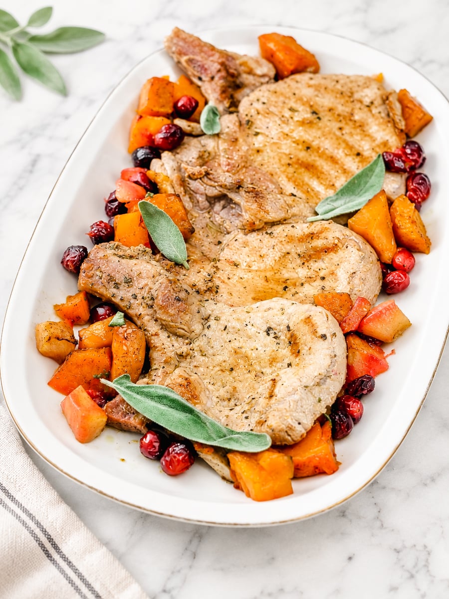 Three cooked pork chops garnished with sage leaves, surrounded by roasted butternut squash and cranberries-this Pork Chops with Pumpkin and Apples-inspired dish is beautifully arranged on a white oval platter atop a marble surface.
