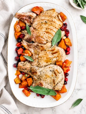Grilled Pork Chops with Pumpkin and Apples are beautifully arranged on a white platter, garnished with fresh sage leaves and surrounded by roasted butternut squash and cranberries. A beige napkin and small bowl are nearby on a marble surface.