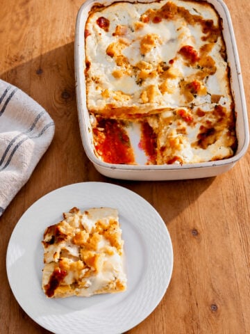 A baked Roasted Butternut Squash and Pumpkin Lasagna with a golden, slightly crispy top in a rectangular dish sits on a wooden table. A square piece is served on a white plate nearby. A striped cloth is also on the table.