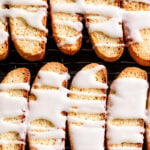 Rows of golden Lemon Biscotti cookies on a cooling rack, drizzled generously with white icing. Thick, uneven stripes of icing accent the bright, citrusy aroma wafting from these crisp, oblong treats.