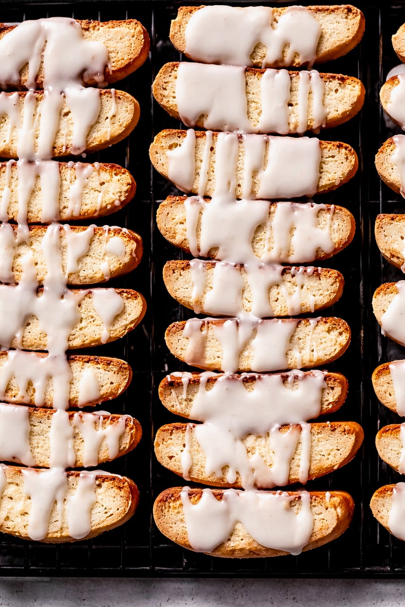 Rows of golden-brown Lemon Biscotti topped with white icing are arranged neatly on a black wire cooling rack. The icing is drizzled unevenly over the crisp cookies.