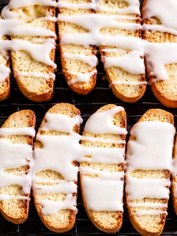 Rows of golden Lemon Biscotti cookies on a cooling rack, drizzled generously with white icing. Thick, uneven stripes of icing accent the bright, citrusy aroma wafting from these crisp, oblong treats.