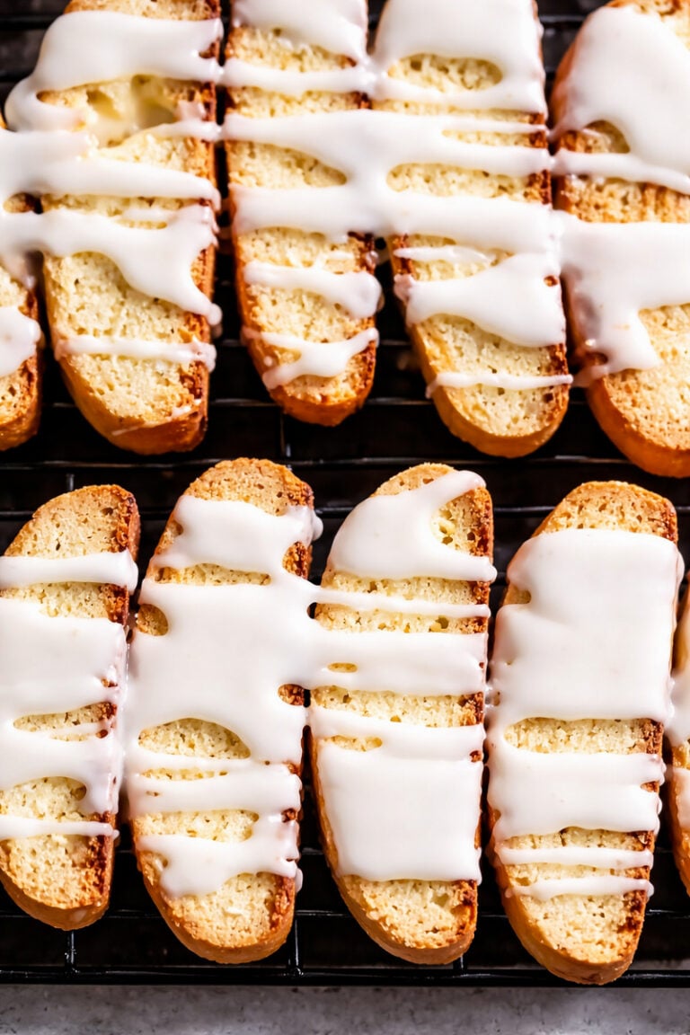 Rows of golden Lemon Biscotti cookies on a cooling rack, drizzled generously with white icing. Thick, uneven stripes of icing accent the bright, citrusy aroma wafting from these crisp, oblong treats.