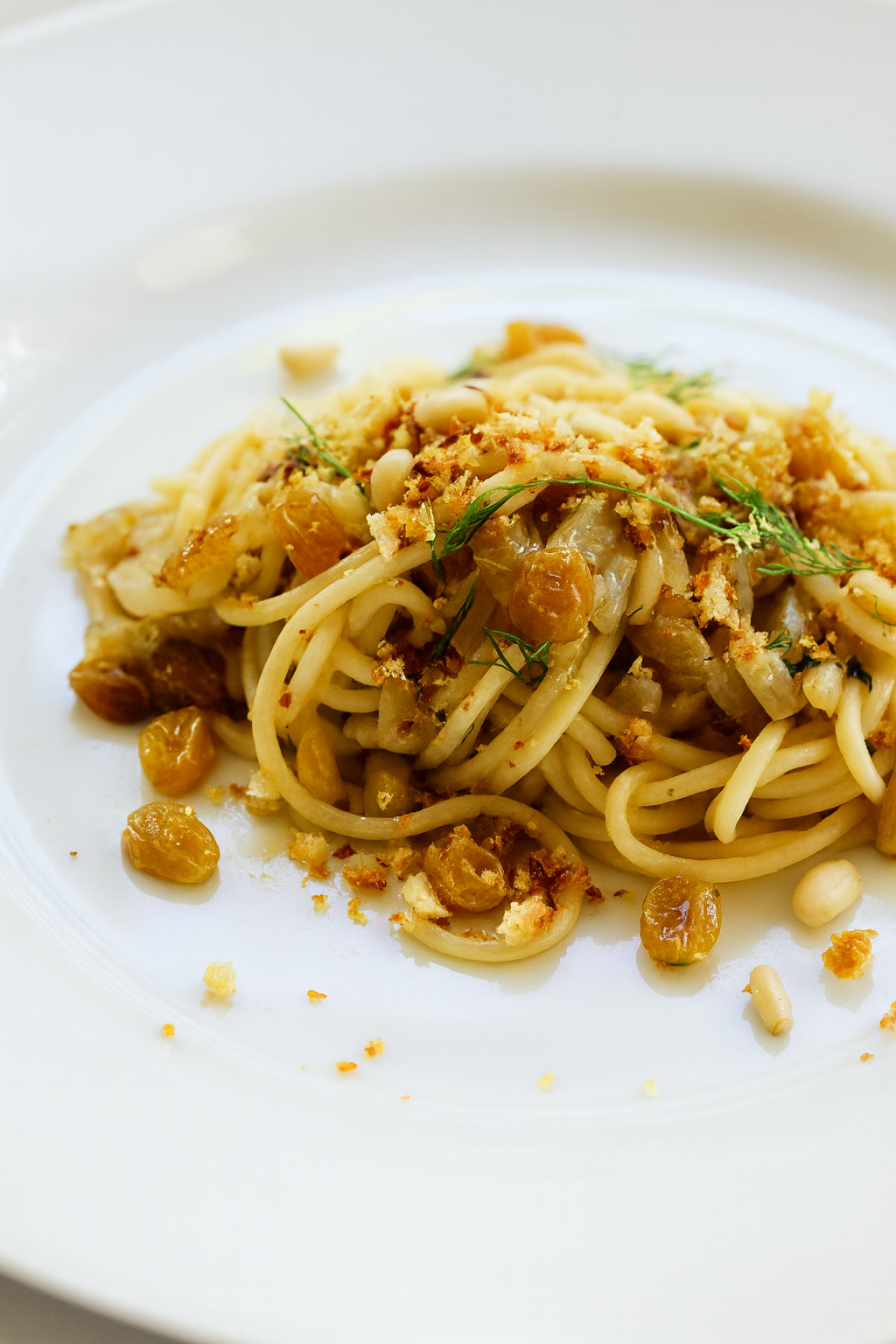 A plate of Pasta con le Sarde featuring spaghetti topped with golden raisins, toasted breadcrumbs, pine nuts, caramelized onions, and fresh herbs, served on a white dish.