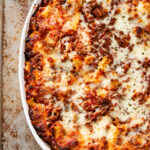 Ziti al Forno baked pasta casserole topped with melted, browned cheese and tomato sauce in a white oval dish on a rustic metal surface.