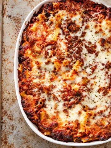 Ziti al Forno baked pasta casserole topped with melted, browned cheese and tomato sauce in a white oval dish on a rustic metal surface.
