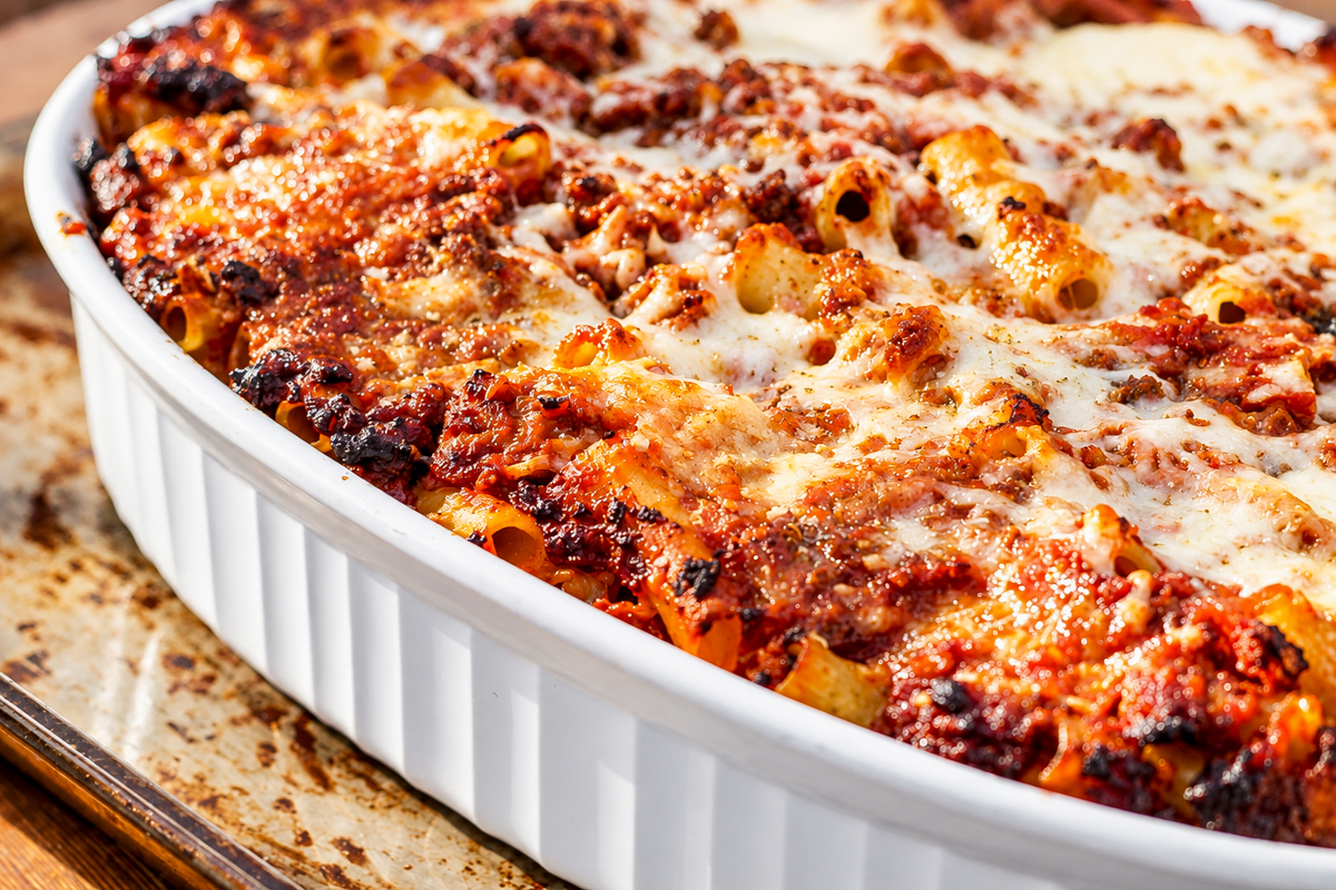 A close-up of a Ziti al Forno casserole in a white dish, showing golden melted cheese, browned edges, and layers of pasta with tomato sauce and meat.