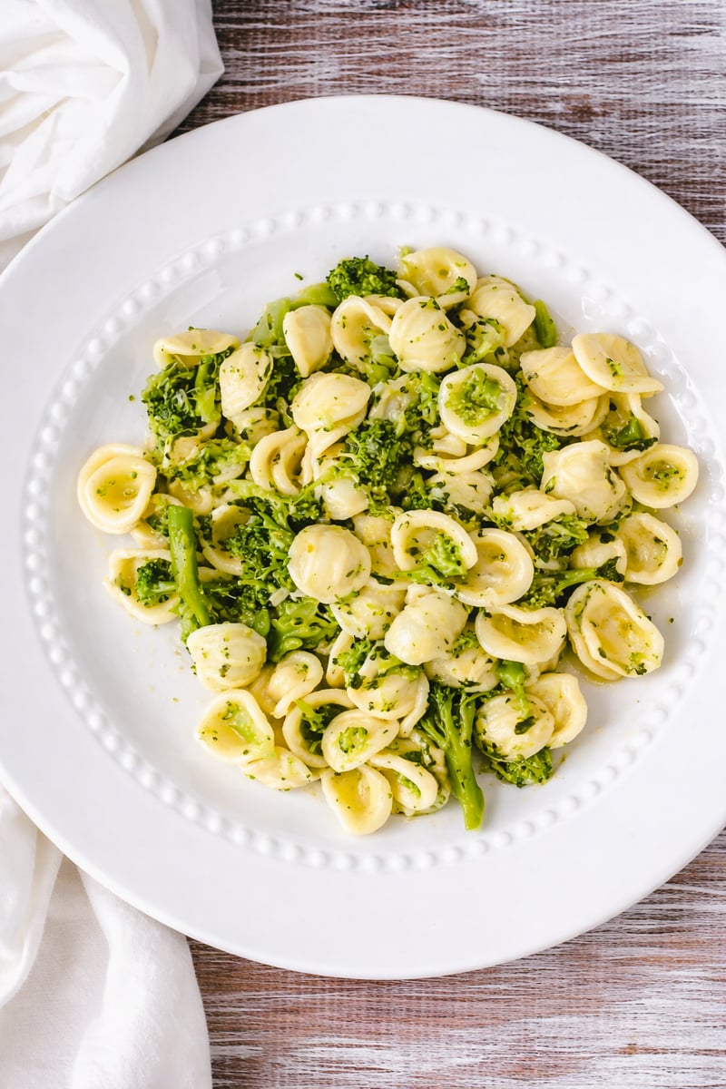 A white plate of Orecchiette Pasta with Broccoli Sauce, garnished with herbs, sits on a rustic wooden table. A white cloth napkin is partially visible beside the plate.