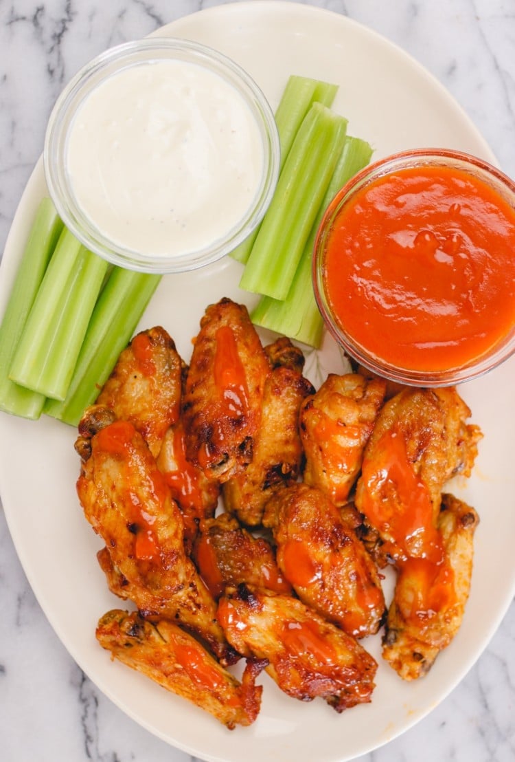 overhead image of buffalo wings with blue cheese dressing, celery sticks, and hot sauce on a white serving platter.