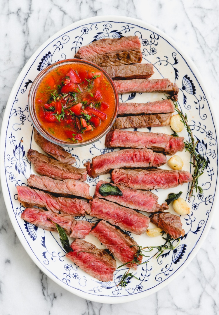 overhead image of sliced steak on a white and blue plate with tomatoes in a glass dish