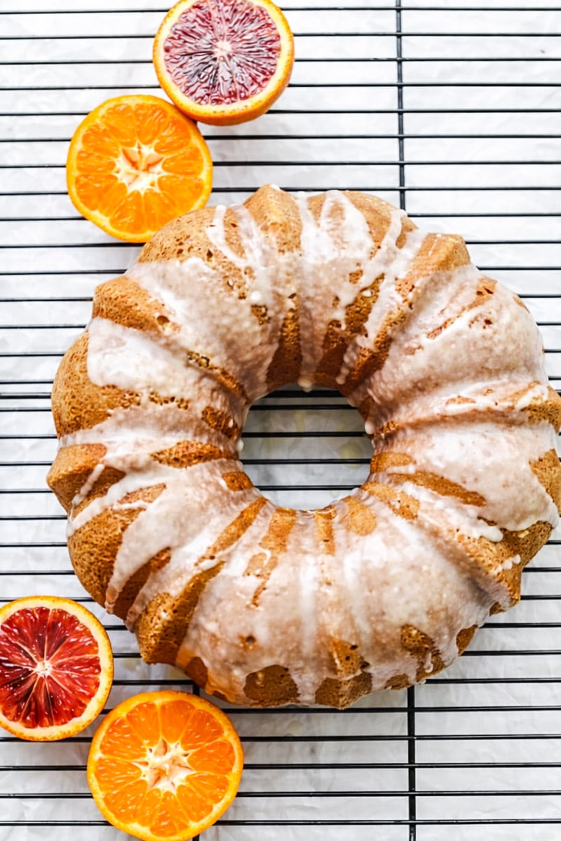 An Orange Bundt Cake with Orange Glaze sits on a cooling rack, surrounded by halved blood oranges and tangerines. The cake is coated in thin white icing, creating a bright and inviting scene.