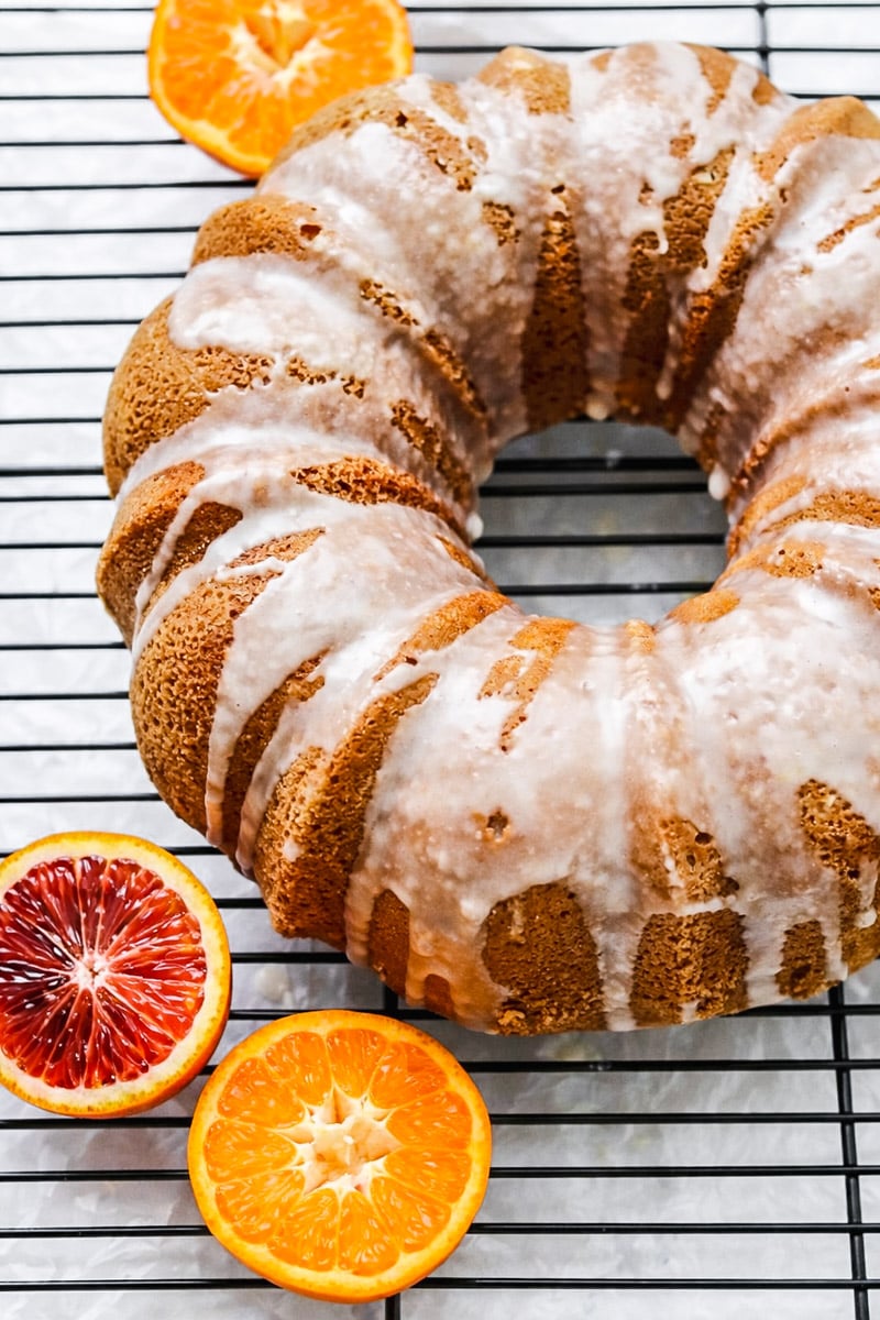 An Orange Bundt Cake with Orange Glaze sits on a cooling rack, surrounded by halved blood oranges and mandarins. The cake features a drizzled white icing atop its golden-brown exterior.