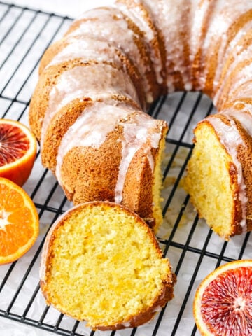 An Orange Bundt Cake with Orange Glaze sits on a black cooling rack, two slices cut to reveal its golden-yellow interior. Fresh, halved blood oranges are arranged beside the cake, which is topped with a light citrusy icing.
