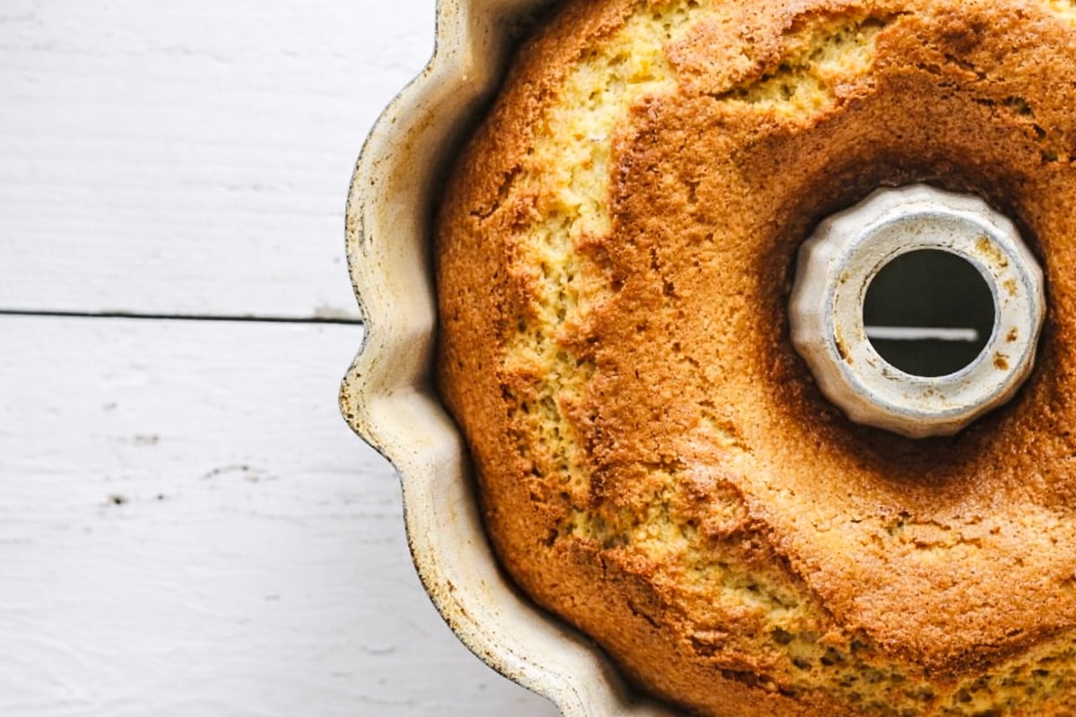 Close-up of a golden-brown Orange Bundt Cake with Orange Glaze in a pan, sitting on a white wooden surface, with half of the cake visible in the frame.