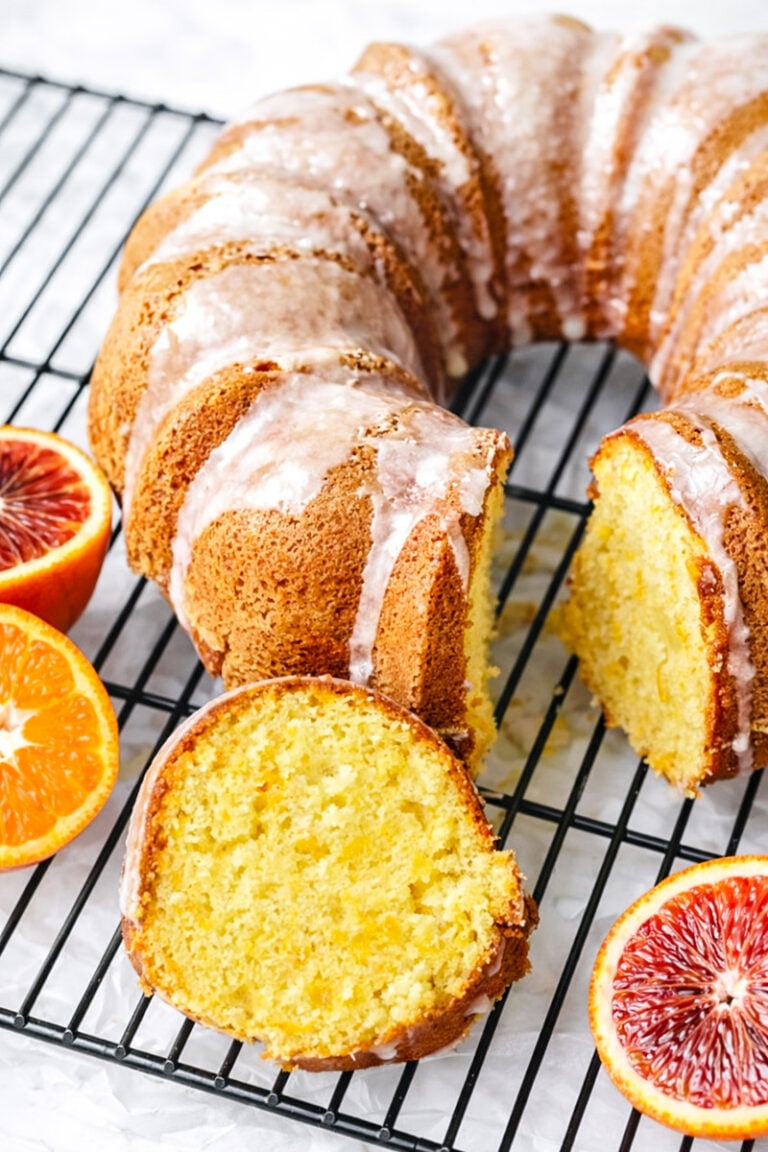 An Orange Bundt Cake with Orange Glaze sits on a black cooling rack, two slices cut to reveal its golden-yellow interior. Fresh, halved blood oranges are arranged beside the cake, which is topped with a light citrusy icing.