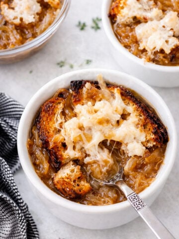 A bowl of Vegan Italian Onion Soup topped with toasted bread and melted cheese, with a spoon resting inside. Another bowl and a small dish are visible in the background, next to a black-and-white striped napkin.