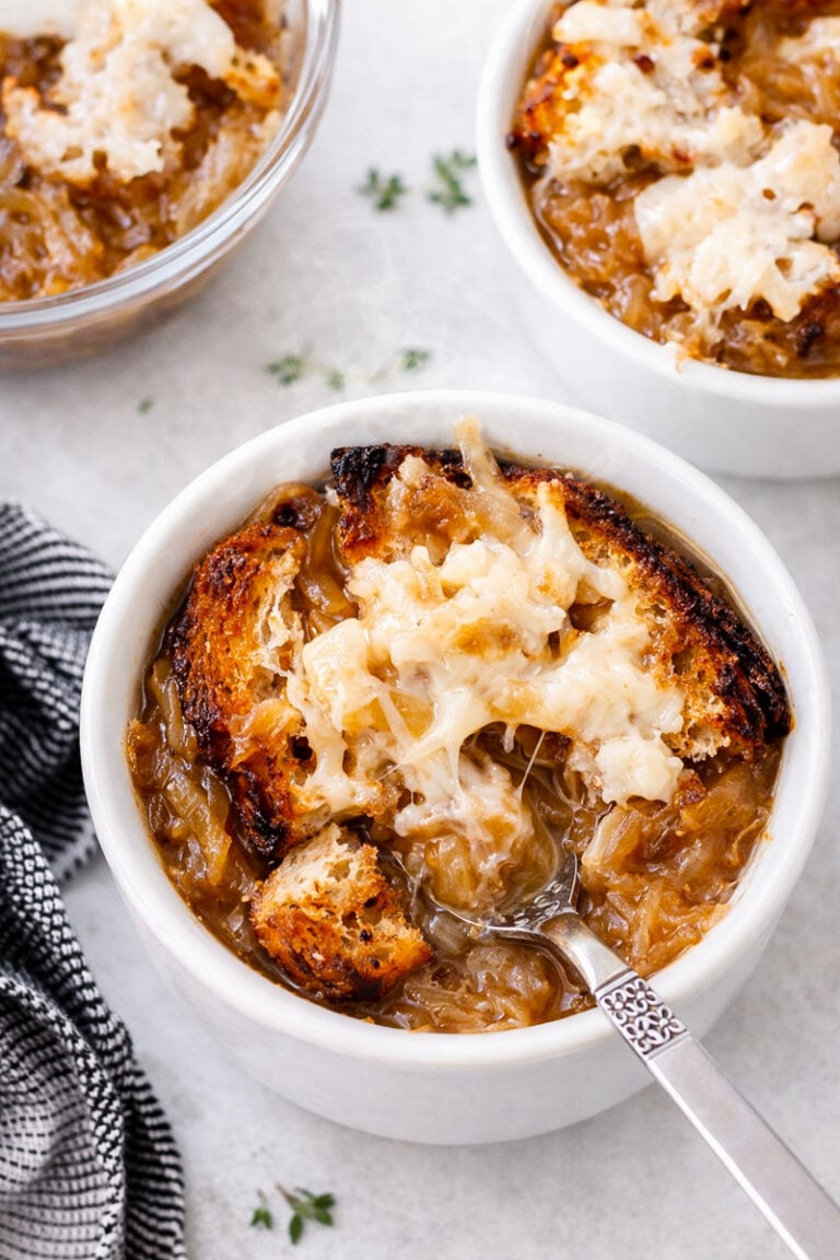 A bowl of Vegan Italian Onion Soup topped with toasted bread and melted cheese, with a spoon resting inside. Another bowl and a small dish are visible in the background, next to a black-and-white striped napkin.