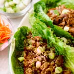 Romaine lettuce leaves filled with seasoned ground meat for chicken lettuce wraps, diced mushrooms, and topped with sliced green onions and sesame seeds, with bowls of chopped green onions, shredded carrots, and sauce nearby.