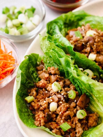Romaine lettuce leaves filled with seasoned ground meat for chicken lettuce wraps, diced mushrooms, and topped with sliced green onions and sesame seeds, with bowls of chopped green onions, shredded carrots, and sauce nearby.