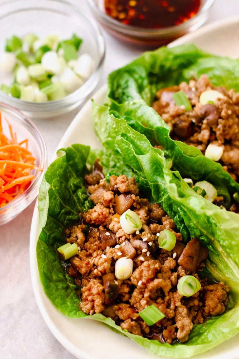 Romaine lettuce leaves filled with seasoned ground meat for chicken lettuce wraps, diced mushrooms, and topped with sliced green onions and sesame seeds, with bowls of chopped green onions, shredded carrots, and sauce nearby.
