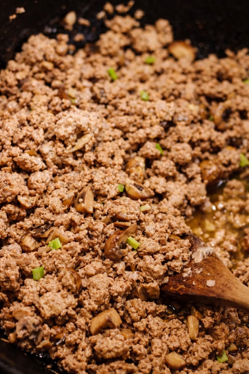 Ground meat for chicken lettuce wraps cooks in a pan with chopped mushrooms and green onions, as a wooden spoon stirs the browned, slightly juicy mixture.