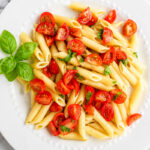 A white plate of vegan cherry tomato pasta with halved cherry tomatoes and chopped basil, garnished with fresh basil leaves, on a marble surface.