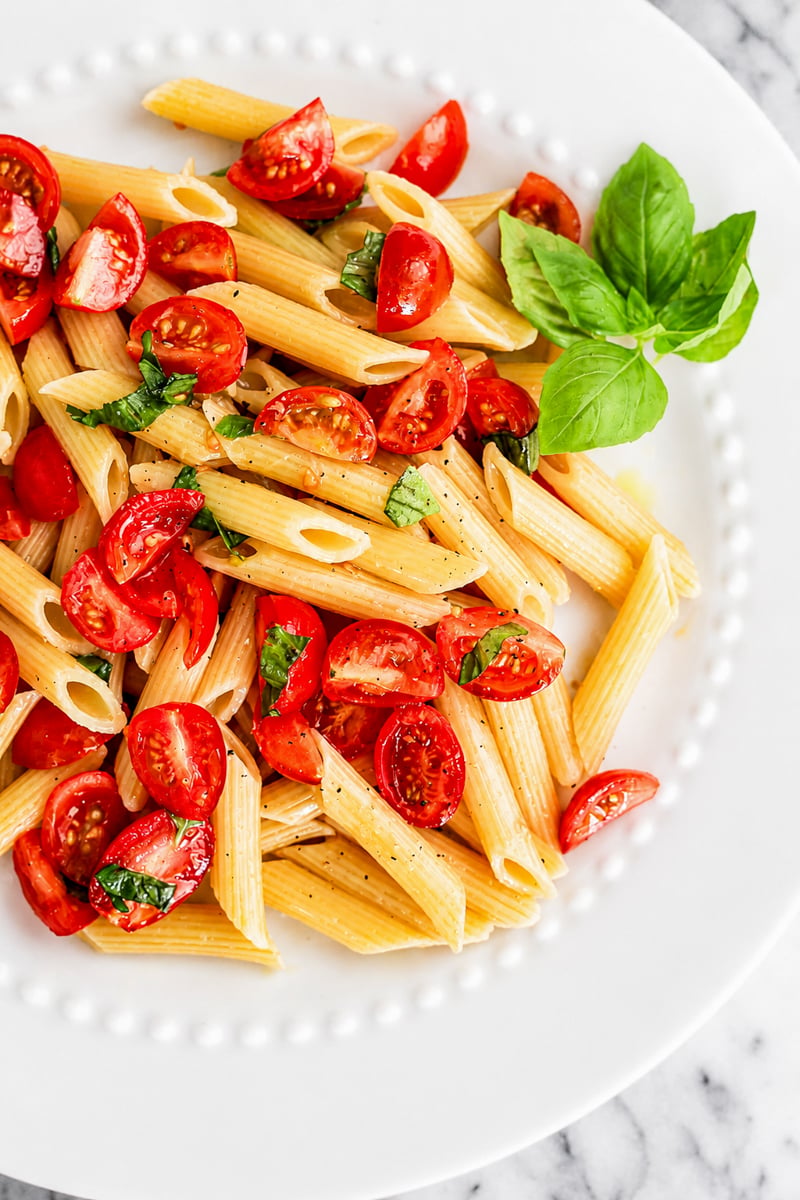 A white plate with vegan cherry tomato pasta, featuring penne pasta, halved cherry tomatoes, fresh basil leaves, and black pepper. The plate is on a marble surface, and a sprig of basil is placed on the side for garnish.