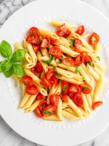 A white plate of vegan cherry tomato pasta with halved cherry tomatoes and chopped basil, garnished with fresh basil leaves, on a marble surface.