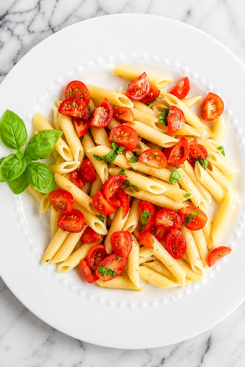 A white plate of vegan cherry tomato pasta with halved cherry tomatoes and chopped basil, garnished with fresh basil leaves, on a marble surface.