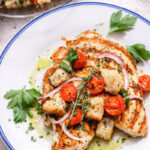 Grilled Chicken Cutlets with a Cherry Tomato Panzanella, topped with croutons, red onion slices, and fresh herbs, served on a white plate with a drizzle of olive oil and parsley. A vibrant salad is visible in the background.
