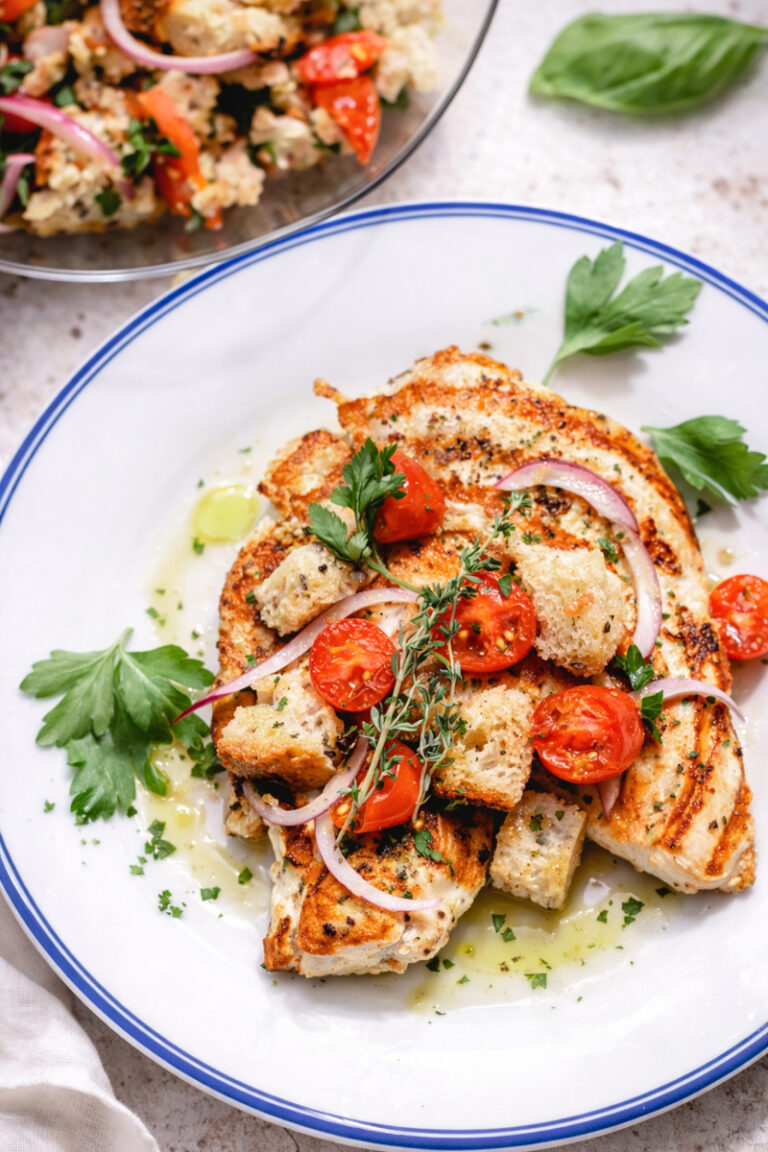 Grilled Chicken Cutlets with a Cherry Tomato Panzanella, topped with croutons, red onion slices, and fresh herbs, served on a white plate with a drizzle of olive oil and parsley. A vibrant salad is visible in the background.