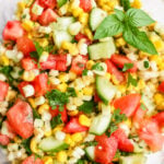 A bowl of Sweet Summer Corn and Cucumber Salad with diced tomatoes, cilantro, and basil leaves, all mixed together in a glass bowl.