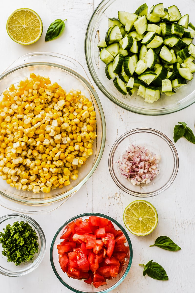 Overhead view of bowls with chopped cucumbers, corn, tomatoes, minced shallots, and herbs on a white surface, perfect for making Sweet Summer Corn and Cucumber Salad. Sliced lime halves and basil leaves are scattered around.