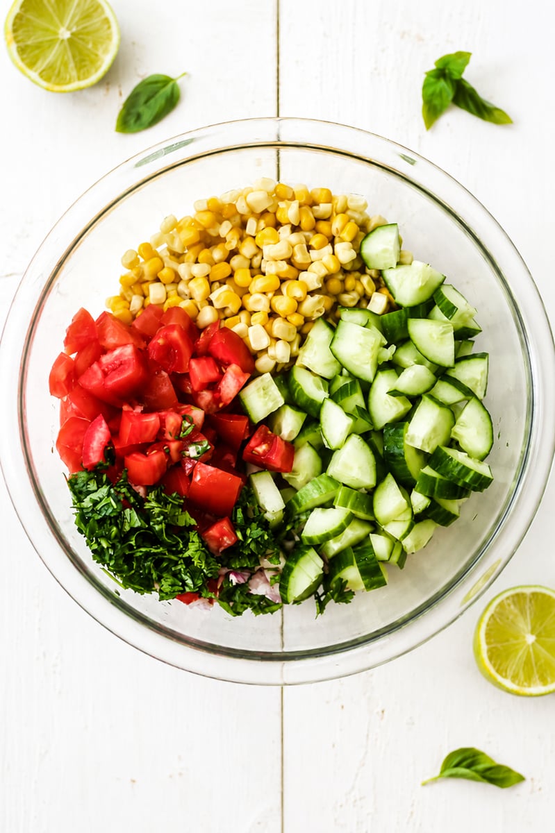 A glass bowl containing Sweet Summer Corn and Cucumber Salad, with tomatoes and herbs arranged in sections. Lime halves and basil leaves are scattered on a white wooden surface around the bowl.