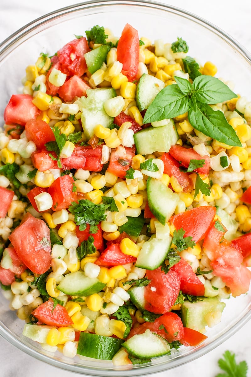 A bowl of Sweet Summer Corn and Cucumber Salad with diced tomatoes, cilantro, and basil leaves, all mixed together in a glass bowl.