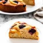 A slice of Gluten-Free Peach and Blackberry Cake, studded with juicy blackberries and peaches, sits on a white plate in the foreground, with the remaining cake on another plate behind it. The scene is set on a marble surface.