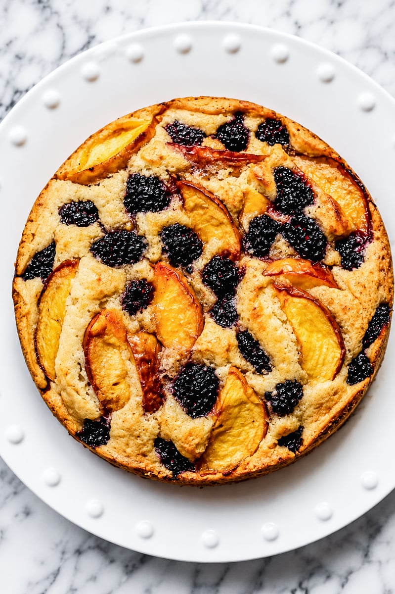 A Gluten-Free Peach and Blackberry Cake, topped with slices of peaches and blackberries, sits on a white, textured plate placed on a marble surface.