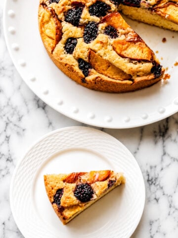 A slice of Gluten-Free Peach and Blackberry Cake, topped with blackberries and peach slices, sits on a small white plate beside the rest of the cake on a larger plate, both displayed on a marble surface.