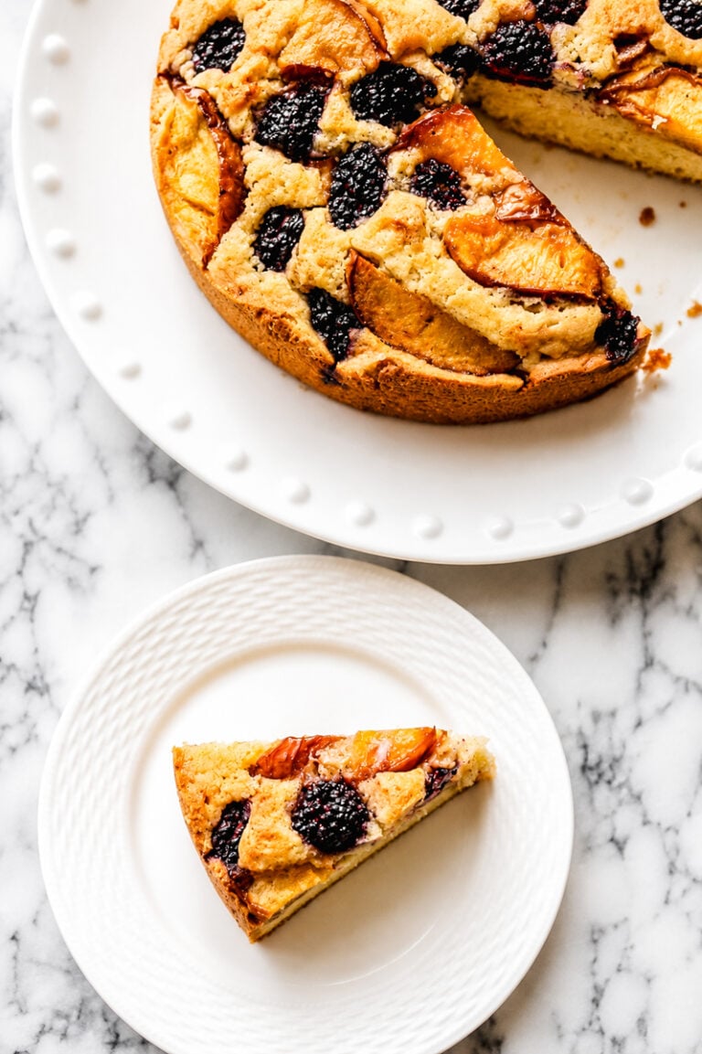 A slice of Gluten-Free Peach and Blackberry Cake, topped with blackberries and peach slices, sits on a small white plate beside the rest of the cake on a larger plate, both displayed on a marble surface.