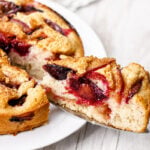 A slice of Easy Italian Plum Cake is being lifted from a larger round cake on a white plate. The cake has a golden crust and visible baked plum slices throughout.