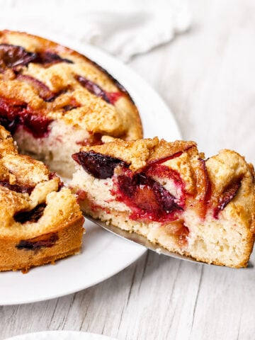 A slice of Easy Italian Plum Cake is being lifted from a larger round cake on a white plate. The cake has a golden crust and visible baked plum slices throughout.